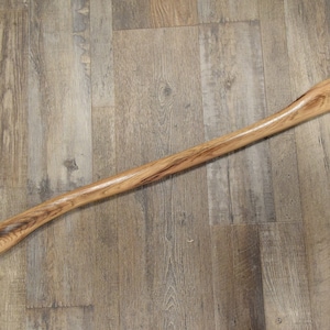 May include: A wooden axe handle with a natural finish. The handle is long and slender with a slight curve at the bottom.