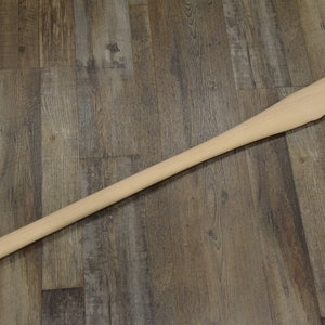 May include: A wooden axe handle with a smooth, light brown finish. The handle is about 36 inches long and has a slightly rounded end.