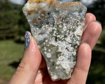 May include: A close-up of a green, white, and brown agate stone with a speckled pattern. The stone is being held in a hand.