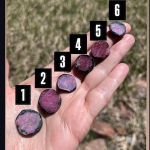 May include: Six small, round, polished gemstones in varying shades of pink and purple are displayed in a hand. Each stone is numbered 1 through 6 in black boxes. The stones have a dark border.