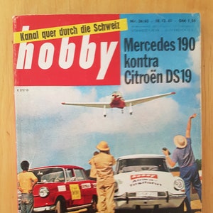 Può includere: Una copertina di rivista vintage con un aereo rosso e bianco che vola sopra un paesaggio desertico. La rivista è intitolata "hobby" e presenta il testo "Mercedes 190 kontra Citroën DS19" e "4. Super-Test mit gekauften Autos: In die Sahara".