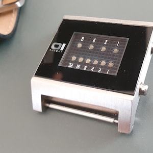 Op de afbeelding: Een zilveren en zwarte digitale horloge met een zwarte wijzerplaat en een zilveren band. De wijzerplaat van het horloge heeft een raster van kleine lampjes die de tijd weergeven. Op de wijzerplaat van het horloge staat ook de tekst "01 THE ONE".