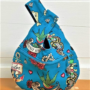 May include: A turquoise blue tote bag with a knot handle. The bag is made of fabric and has a zipper closure. The fabric has a pattern of various tattoos, including hearts, roses, anchors, swallows, and snakes. The text "Forever Love" is repeated on the bag.
