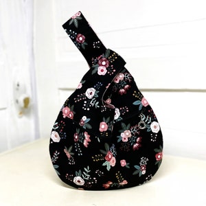 May include: A black fabric tote bag with a floral pattern in shades of pink, white, and green. The bag has a knotted handle and a small metal loop on the side.