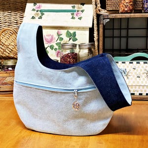 May include: A light blue denim tote bag with a dark blue denim strap. The bag has a zipper closure and a decorative metal charm.