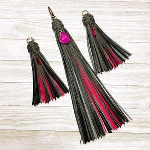 May include: Three leather tassels in varying lengths featuring dark gray and bright pink fringe strands. Each tassel is topped with a braided leather cap and a metal ring, accented by a small, dangling, polished pink or red stone charm.