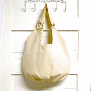May include: A large, off-white canvas tote bag with a floral print lining. The bag has a gathered bottom and is hanging from a white hook.