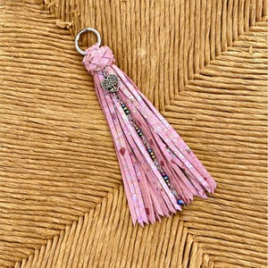 May include: A pink tassel keychain featuring a metallic silver split ring, a braided top knot, and a silver-toned heart-shaped tree charm. The tassel consists of multiple thin, iridescent pink strips accented by a single strand of small, multicolored beads.