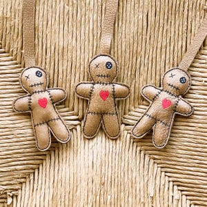May include: Three tan doll-shaped ornaments made of textured material, each featuring a red heart stitched on the chest, black stitched mouth and limbs, and two mismatched eyes, one being a black button and the other a black stitched X.