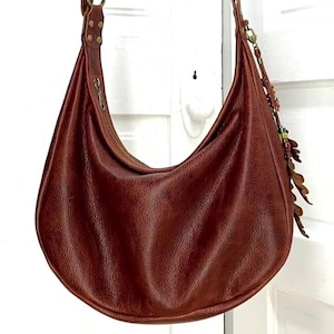 May include: A crescent-shaped hobo bag crafted from textured brown leather. The bag features a single shoulder strap attached with brass-toned rivets, a top zipper closure, and a decorative charm with leaf-shaped cutouts and colorful beads.