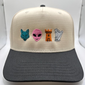 May include: A beige and black baseball cap with embroidered patches. The patches include a teal fox, a pink alien head, an orange castle, and a silver harp. The cap has a structured fit with a curved brim.