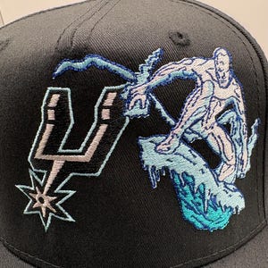 May include: Black baseball cap featuring an embroidered design. The design includes the San Antonio Spurs logo and a stylized figure riding a wave. The figure and wave are in shades of blue and white.