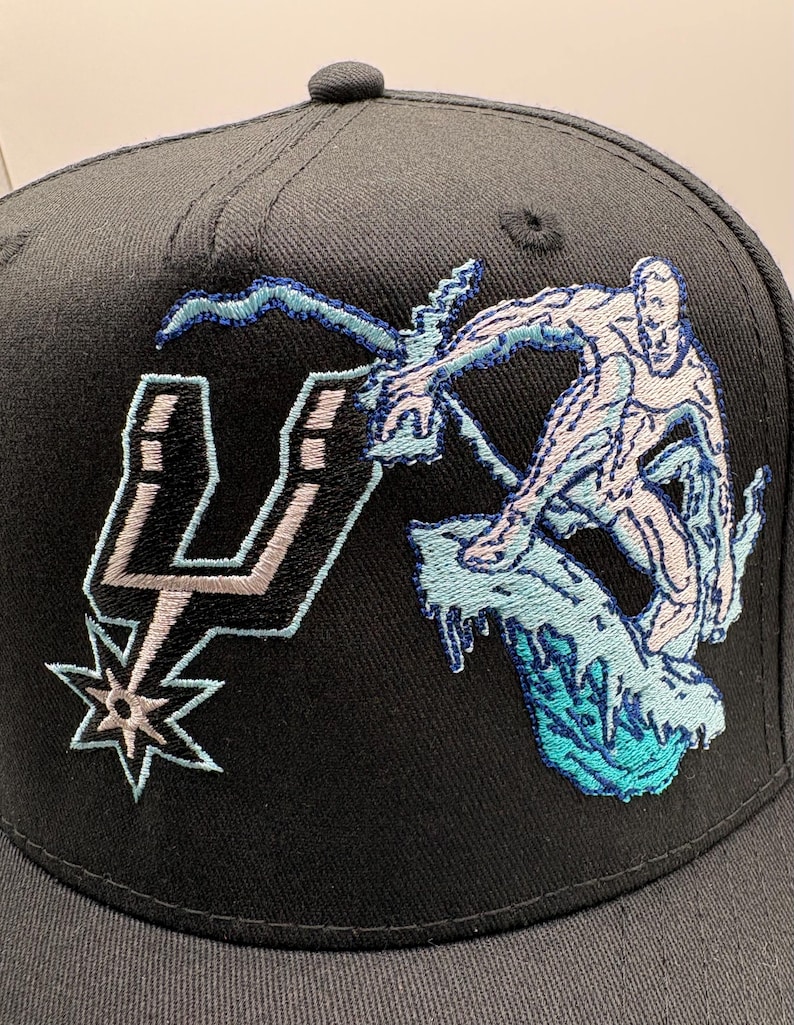May include: Black baseball cap featuring an embroidered design. The design includes the San Antonio Spurs logo and a stylized figure riding a wave. The figure and wave are in shades of blue and white.