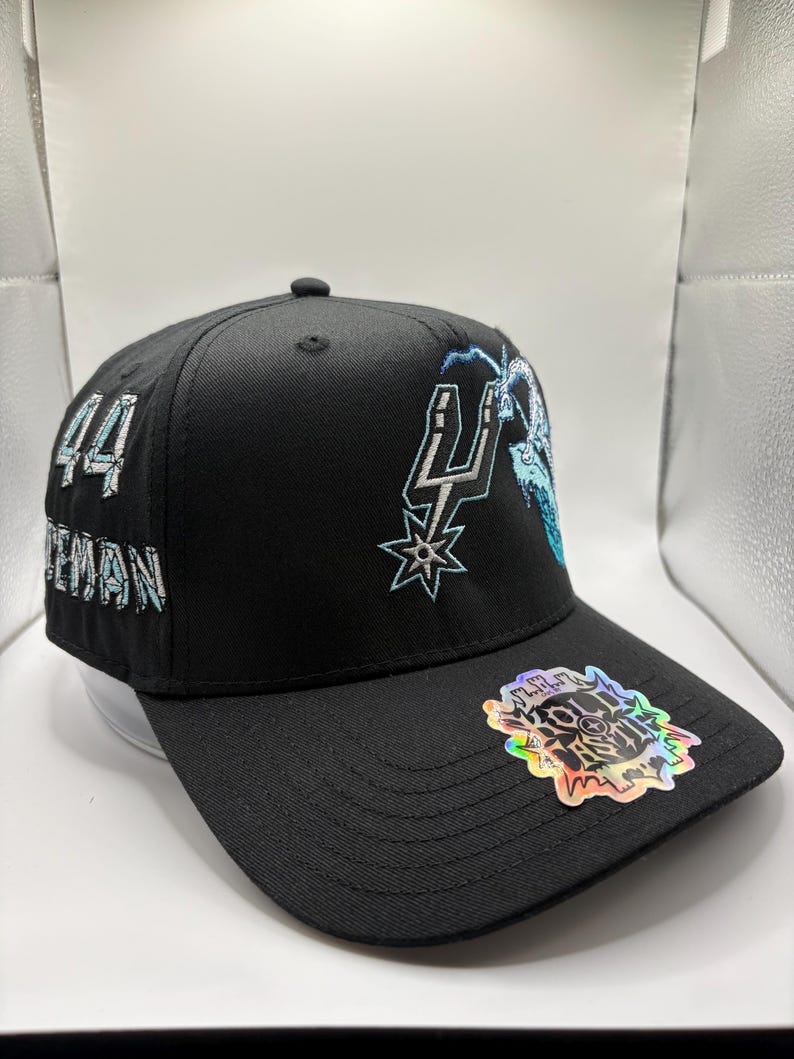 May include: Black baseball cap with embroidered team logo and the number "44" on the side. The cap features a holographic sticker on the brim. The team logo is in silver and light blue.