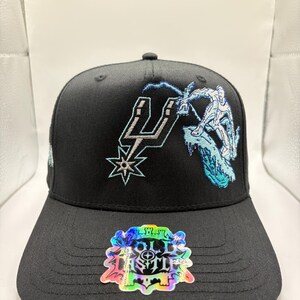 May include: Black baseball cap featuring the San Antonio Spurs logo and a graphic of a figure snowboarding. The cap has a flat brim and a sticker with the text "OLD CASTLE". The embroidery is in white and light blue.