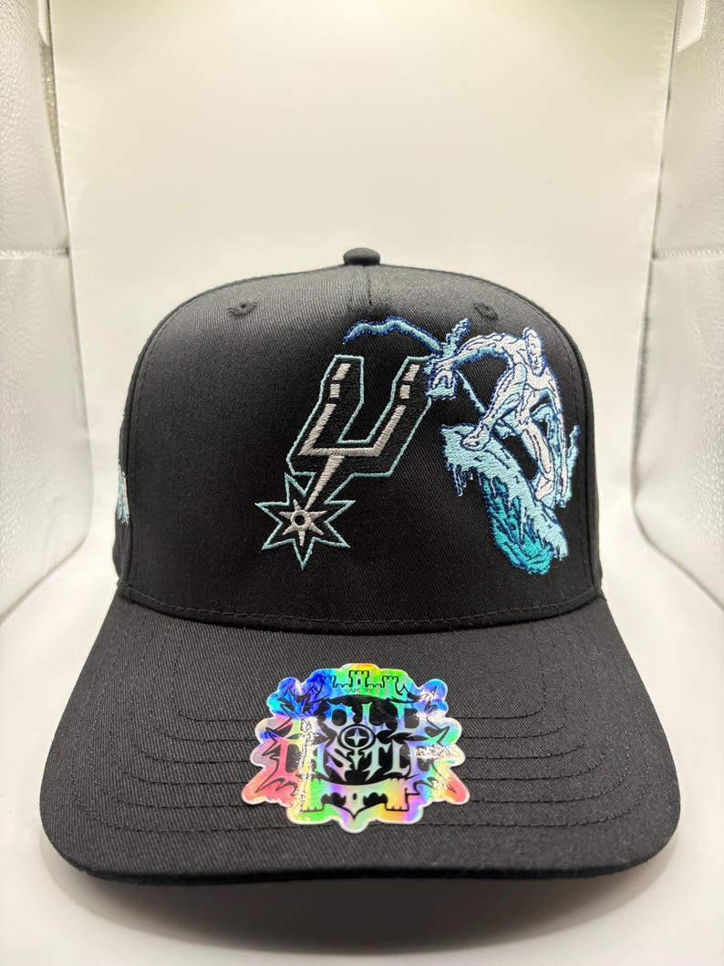 May include: Black baseball cap featuring the San Antonio Spurs logo and a graphic of a figure snowboarding. The cap has a flat brim and a sticker with the text "OLD CASTLE". The embroidery is in white and light blue.