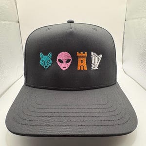 May include: A charcoal gray baseball cap featuring four embroidered icons centered on the front panel: a teal fox head, a pink alien face, an orange castle tower, and a white harp. The cap has a structured crown and a flat, stitched brim.