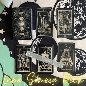 Puede incluir: Una baraja de tarot negra con ilustraciones doradas, con cartas como "El Mago" y "Reina de Oros". Una varita de selenita descansa sobre las cartas. La baraja se muestra sobre una tela con estampado celestial en blanco y negro. El texto "Luna Somnia Deck" es visible.