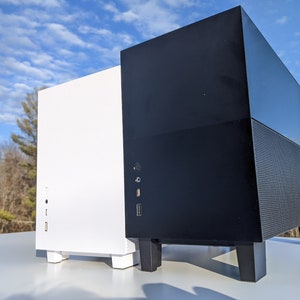 May include: Two computer cases, one black and one white, are shown side-by-side on a table. Both cases have a similar design with a rectangular shape and a front panel with ports. The black case has a textured bottom panel.
