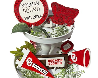 Puede incluir: Una bandeja de servir blanca de dos niveles con una variedad de decoraciones con el tema de la Universidad de Oklahoma. Las decoraciones incluyen un letrero redondo que dice "The University of Oklahoma Norman Bound Fall 2024 1890", un letrero de madera que dice "Boomer Sooner", un banderín rojo y blanco que dice "Sooners", un carro rojo con la palabra "Sooners" en él, y un megáfono rojo.