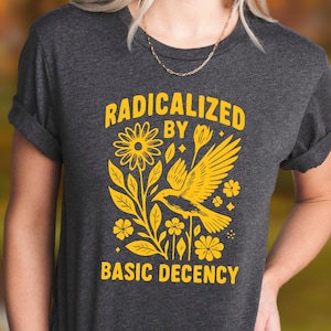 May include: Dark grey t-shirt with the text "RADICALIZED BY BASIC DECENCY" in yellow. The graphic features a yellow bird, flowers, and leaves. The shirt has rolled-up sleeves.