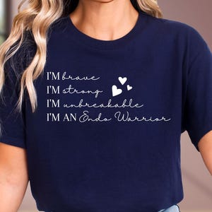 May include: Navy blue t-shirt with white script text that reads "I'M brave, I'M strong, I'M unbreakable, I'M AN Endo Warrior." The shirt also features three small white heart graphics. The shirt is made of a soft material.