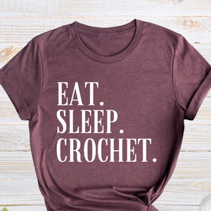May include: A maroon t-shirt with the words "EAT. SLEEP. CROCHET." in white block letters. The shirt is knotted at the bottom and is next to a small succulent plant in a pot and a pair of white sneakers.