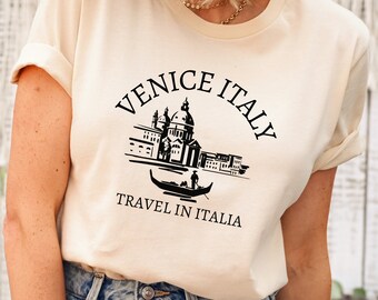 May include: A cream colored t-shirt with a black graphic of a gondola in Venice, Italy. The text "Venice Italy" is in a black arc above the gondola and the text "Travel in Italia" is below the gondola.