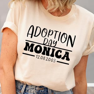 May include: A cream-coloured t-shirt with the words "ADOPTION DAY MONICA 12.08.2003" printed in black. The text is centered and features a bold, stylised font. The shirt has short sleeves and a crew neck.