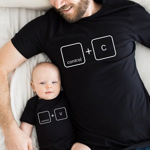 May include: A father and child wearing matching black t-shirts with white computer keyboard key graphics. The father's shirt says "control + c" and the child's shirt says "control + v".