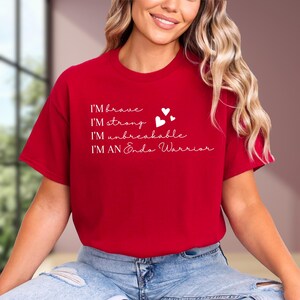 May include: A red t-shirt with white text that reads "I'M brave, I'M strong, I'M unbreakable, I'M AN Endo Warrior." Two white hearts are next to the words "I'M strong." The shirt is made of a soft material.