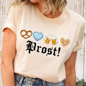 A woman wearing a white t-shirt with a graphic design featuring a beer mug, pretzel, heart, and the word "Prost!" printed on it.