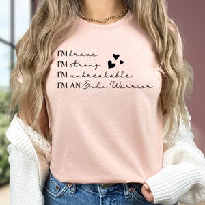 May include: A light pink t-shirt with the phrases "I'm brave, I'm strong, I'm unbreakable, I'm an Endo Warrior" in black script. Three small black hearts are printed next to "I'm strong". The shirt is worn by a person.