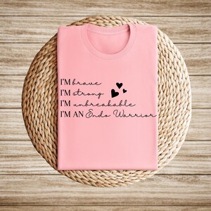 May include: A light pink t-shirt with the words "I'M brave, I'M strong, I'M unbreakable, I'M AN Endo Warrior" in black script. Three small black hearts are next to the words "I'M strong" and "I'M unbreakable". The shirt is folded and placed on a woven, circular surface.