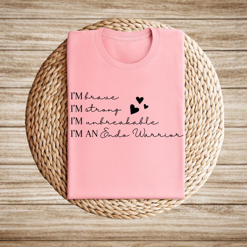 May include: A light pink t-shirt with the words "I'M brave, I'M strong, I'M unbreakable, I'M AN Endo Warrior" in black script. Three small black hearts are next to the words "I'M strong" and "I'M unbreakable". The shirt is folded and placed on a woven, circular surface.