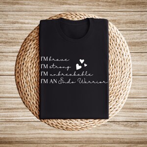 May include: Black t-shirt with white text: "I'M brave, I'M strong, I'M unbreakable, I'M AN Endo Warrior." Three small white hearts are also printed on the shirt. The shirt is displayed on a round, woven, light brown surface.