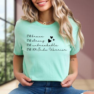 May include: A light green t-shirt with the text "I'M brave, I'M strong, I'M unbreakable, I'M AN Endo Warrior" in black script. The shirt also features three black heart graphics. The model is wearing blue jeans.