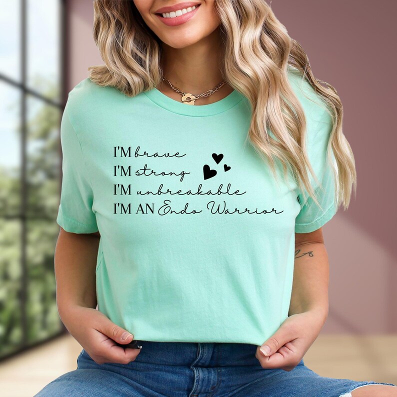 May include: A light green t-shirt with the text "I'M brave, I'M strong, I'M unbreakable, I'M AN Endo Warrior" in black script. The shirt also features three black heart graphics. The model is wearing blue jeans.