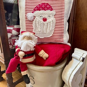 May include: A decorative Christmas pillow with a Santa Claus face on a red and white striped background. A Santa Claus doll sits on a red sack. The pillow and doll are on a cream-colored ceramic pot.