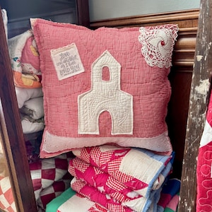 Può includere: Un cuscino a quadretti rossi e bianchi con un'applicazione a forma di chiesa bianca e un centrino di pizzo. Il cuscino ha un'etichetta con la scritta "Love one another as I have Loved You". Sono visibili trapunte impilate in vari modelli e colori.