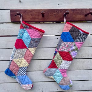 May include: Two quilted Christmas stockings made from a patchwork of various fabric scraps in colours including red, pink, blue, yellow, and white. Each stocking features a red fabric trim at the top opening and a white hanging loop for display.
