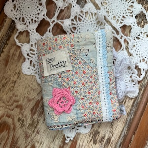 May include: A small blue and white patchwork fabric pouch with a light blue crocheted trim. The pouch has a pink crocheted flower and a label that says "Sew Pretty".