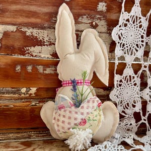 May include: A white fabric bunny with a pink ribbon and lavender flowers. The bunny is holding a heart-shaped patchwork fabric with floral and plaid patterns. The bunny has a white pom-pom tail. The bunny is sitting on a wooden surface with a white crocheted doily.