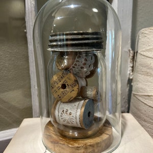 Op de afbeelding: Een glazen stolp met een houten voet, die een glazen pot met een metalen deksel bevat, gevuld met spoelen garen, kant en een blad muziek.