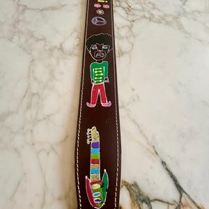 Könnte beinhalten: Ein brauner Gitarrengurt aus Leder mit weißen Nähten und farbenfrohen Illustrationen. Der Gurt zeigt eine Cartoonfigur, eine Regenbogen-Gitarre und den Namen "Jimi" in gelben Buchstaben sowie Friedenszeichen und Blumen.