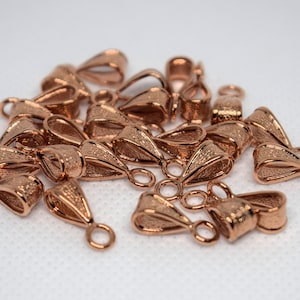 Op de afbeelding: Een verzameling roségouden hangerbeugels. Elke beugel heeft een getextureerd, druppelvormig ontwerp met een kleine lus aan de onderkant voor het bevestigen van een hanger en een grotere lus aan de bovenkant voor een ketting. Ze liggen op een witte ondergrond.