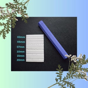 May include: A white square tile sheet with different sized squares, labelled with measurements in millimetres: 03mm, 05mm, 07mm, 10mm, 15mm, and 20mm. A purple ruler is lying on a black surface.