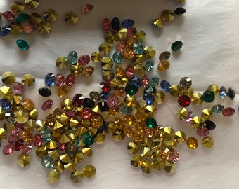 May include: Assortment of colorful rhinestones in various shades of blue, green, red, pink, and gold. The rhinestones are scattered on a white surface.