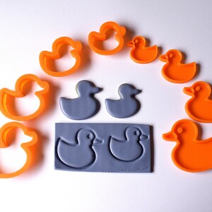 Puede incluir: Un juego de nueve cortadores de galletas de patos de goma, incluyendo dos moldes de silicona grises. Los cortadores son de color naranja y presentan una variedad de diseños de patos.