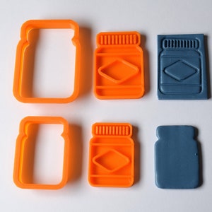 May include: Two orange plastic cookie cutters shaped like jars with lids, and two blue plastic cookie cutters shaped like jars with lids.